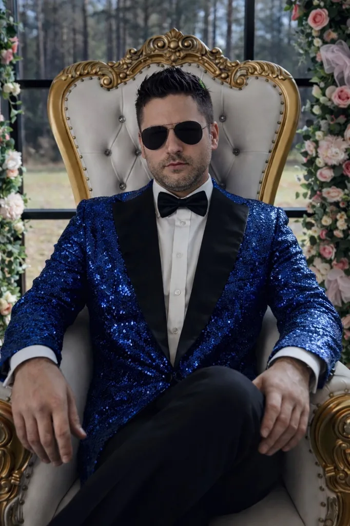 DJ Rayman seated in a white and gold chair in a blue sequin tuxedo jacket, framed by wedding florals